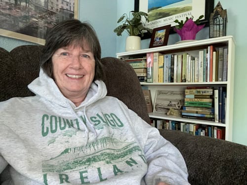 Reviewer smiling while wearing the light gray County Sligo Hoodie while sitting in a cozy chair.
