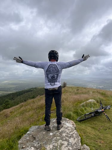 Customer wearing the silver Reaper Long Sleeve Jersey with arms outstretched on a scenic hilltop, showing the back graphic.
