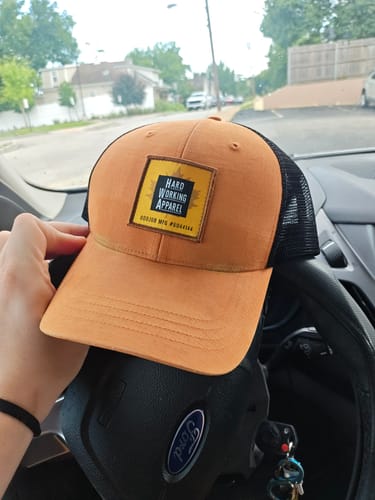 Reviewer's hand holding a tan and black trucker hat over a car's steering wheel.