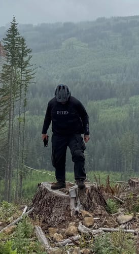 Reviewer wearing the black Highroller Cargo Trousers while standing on a tree stump in a forested area.