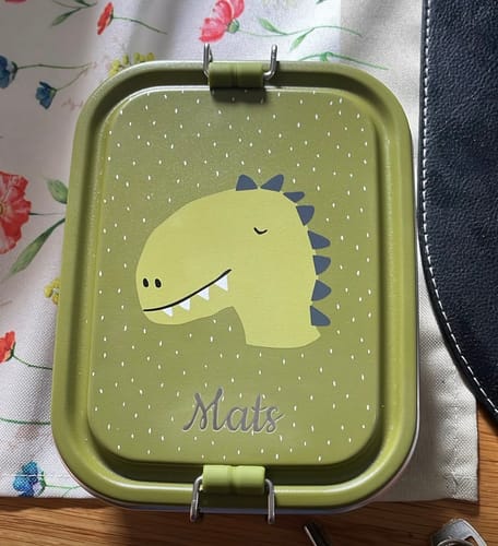 Kunde zeigt die personalisierte grüne trixie Brotdose mit Dinosaurier-Motiv und dem Namen 'Mats'.