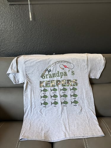 A customer's light grey 'Grandpa's Keepers' fishing shirt spread out on a couch, displaying the personalized fish graphic.