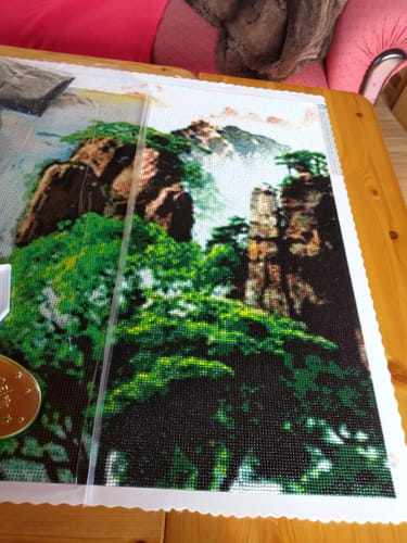 Kundenfoto des Diamond Paintings „Berge Wasserfall-Bäume“, das auf einem Holztisch liegt und sich in Arbeit befindet.