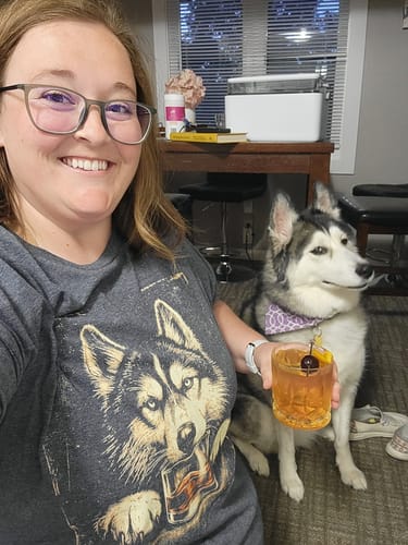 Reviewer wearing the gray Whiskey Husky Grunge T-Shirt while holding a drink and sitting next to a husky.
