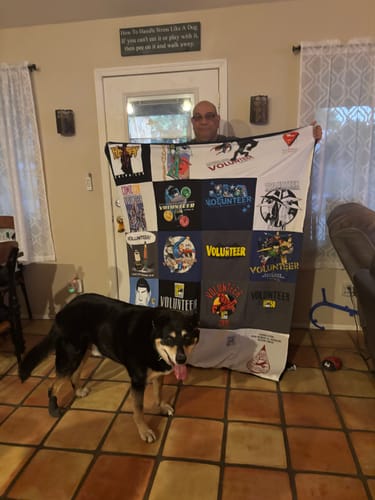 Customer holding up their large Project Repat T-Shirt Quilt made from Comic-Con volunteer shirts, with a dog standing in front.