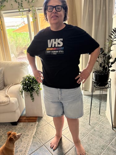 A reviewer stands in their living room wearing the black 'Still Human' t-shirt with light-wash denim shorts.