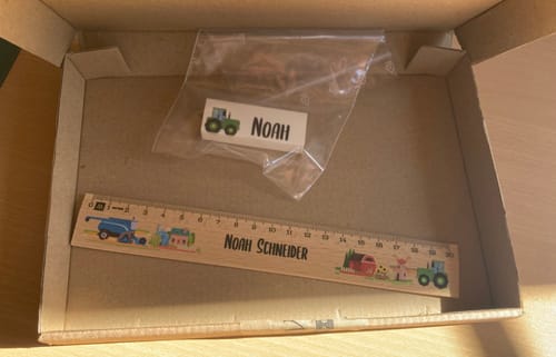 Rezensent zeigt das personalisierte Holzlineal mit bunten Bauernhof-Motiven in der offenen Versandbox.