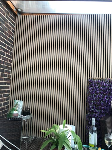Customer's light-colored Acoustic Slatted Wall Panels installed vertically on a covered patio wall next to brick.