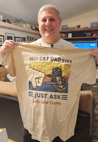 Customer holding up a personalized beige 'Best Cat Dad Ever' shirt with a graphic of two cats and the names Lucy and Candy.