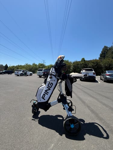 A customer's white Cybercart Push+ holds a golf bag in a sunny parking lot.