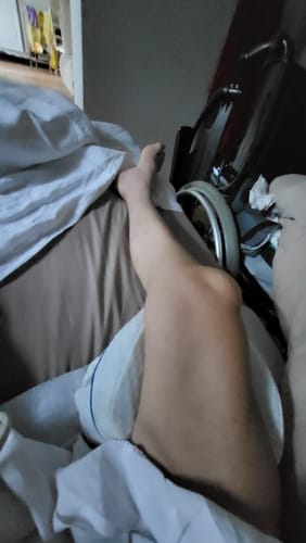 Client utilisant le coussin orthopédique Cellsius pour soutenir sa jambe dans un lit, avec un fauteuil roulant en arrière-plan.