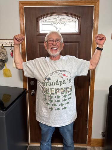 A happy customer wearing the personalized 'Grandpa's Keepers' fishing shirt, flexing his arms indoors.