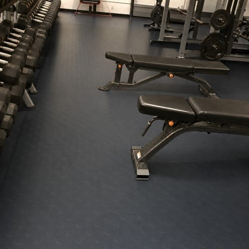 Reviewer's photo of the dark Studded Dot Penny Rubber Flooring installed in a gym, with weight benches on top.