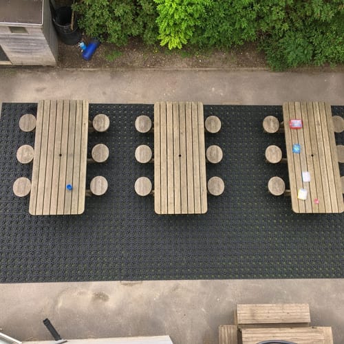 Customer's overhead view of black Rubber Grass Mats installed under three wooden picnic tables on a patio.
