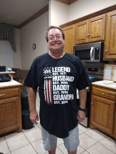 A customer smiling while wearing the black personalized 'Proud Grandpa Legend' shirt in a kitchen.