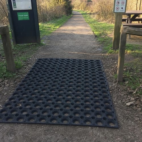 A customer's black Rubber Grass Mat installed on an uneven dirt path at the entrance to a park trail.