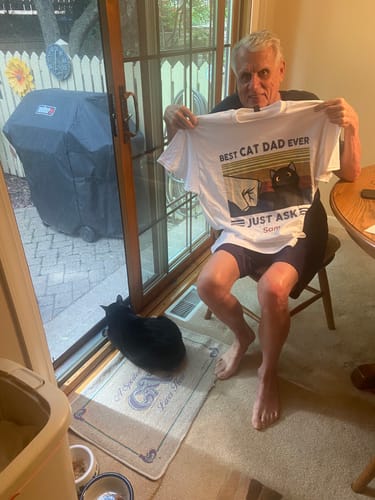 Customer holding his personalized white 'Best Cat Dad Ever' shirt, with his black cat sitting on the floor nearby.