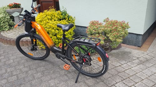 Reviewer's Lankeleisi MG600 Lite e-bike with custom orange grips and pedals, parked on a paved driveway next to bushes.