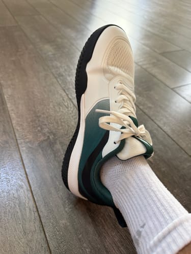 A customer's foot wearing the white and green CUURT Muuv court shoe on a wood floor.