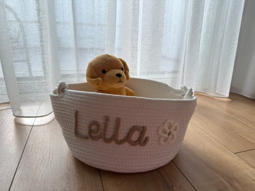 Customer's personalized white woven basket with the name 'Leila' and a flower icon, holding a plush dog on a wood floor.