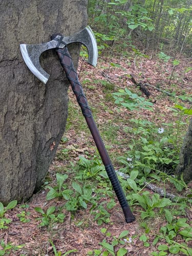 Customer's Viking Double-Edged Axe 'Havamal' with a dark leather-wrapped handle leaning against a tree outdoors.