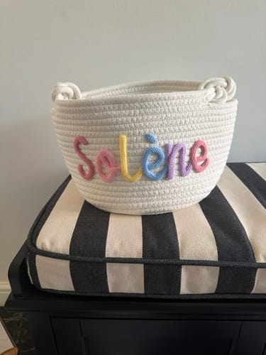 Reviewer's personalized woven basket with the name "Solène" written in colorful pastel yarn, sitting on a striped cushion.