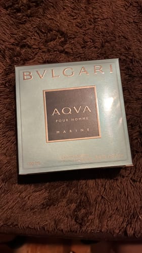 Cliente mostrando la caja de su perfume BVLGARI AQVA MARINE Pour Homme sobre una superficie afelpada.