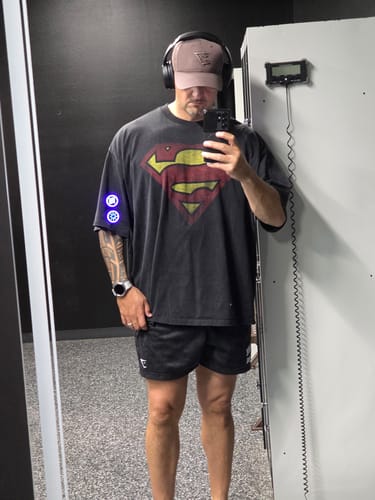 Reviewer in a gym wearing the oversized Superman Huge Tees, showing its dark color and front logo.
