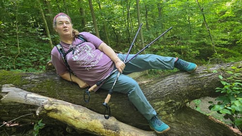 Reviewer playfully resting on a log in a forest while wearing the purple Bad Knees T-shirt.