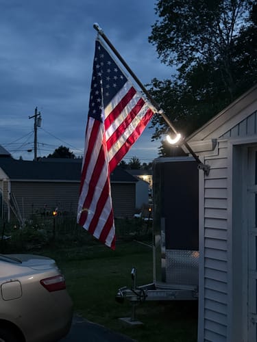 Customer's Prestige House Flag Pole mounted to a garage, with a light illuminating the American flag at dusk.