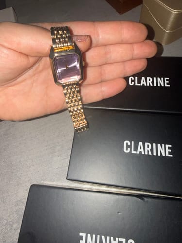Reviewer's hand holding the CLAIRE DE MAINS watch, showing its fancy gold-colored band and purple rectangular face.