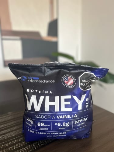 Un cliente muestra el empaque azul de su Proteína Whey sabor Vainilla sobre una mesa.