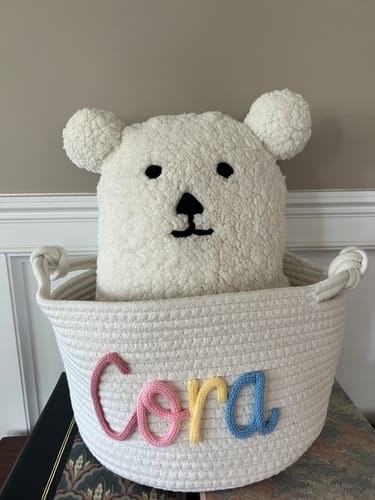 Customer's personalized white rope basket with the name "Cora" in colorful yarn, holding a teddy bear.