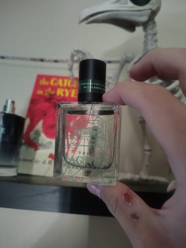 A reviewer's hand holding the clear, square bottle of Jon and Martin perfume, showing its green etched design and black cap.