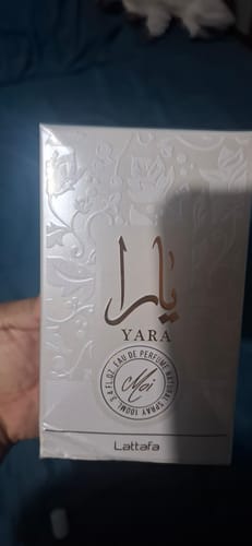 Un cliente sostiene la caja blanca y sellada del perfume Lattafa Yara Moi sobre una superficie azul.