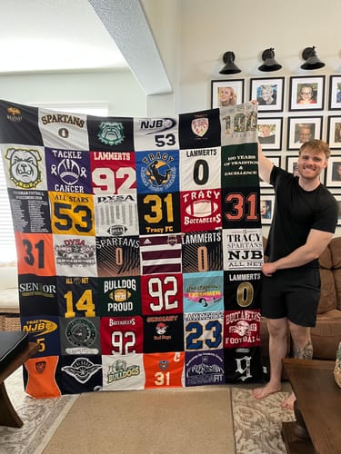 Reviewer holding up a large Project Repat T-Shirt Quilt made from various sports jerseys in a living room.