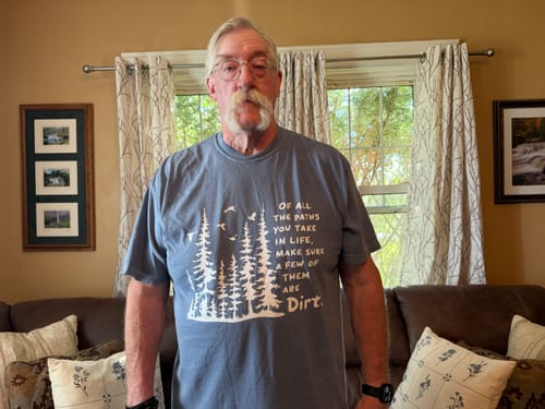 A reviewer wearing the blue-grey Paths In Life T-shirt, showing its relaxed fit and outdoor-themed graphic.