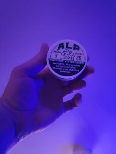 Customer's hand holding a round, white tin of Mountain Wintergreen Nicotine Pouches in purple light.