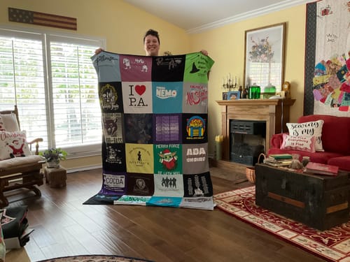 Customer holding up their large, colorful Project Repat T-Shirt Quilt in a living room, showing the various shirts.