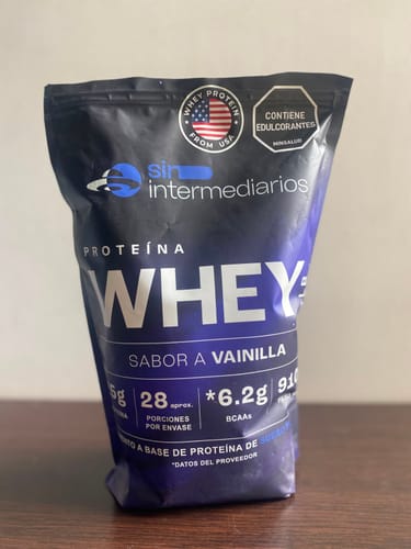 Foto de un cliente del empaque azul de la Proteína Whey sabor Vainilla sobre una mesa.