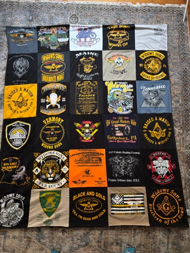 Reviewer's large Project Repat T-Shirt Quilt made from many graphic tees with black sashing, laid out on a patterned rug.