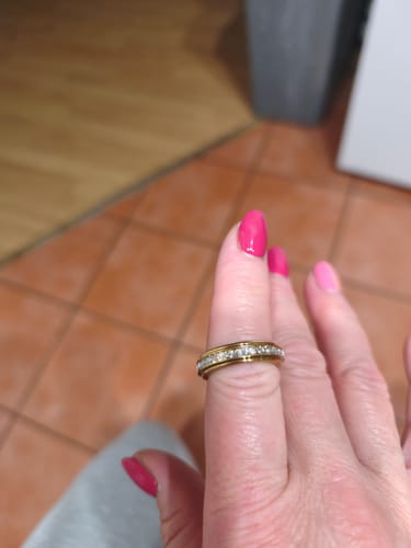 Kliento ranka, mūvinti aukso spalvos Tylos žiedą „Calmy“ su blizgiais akmenukais ant rožinai lakuoto nago.