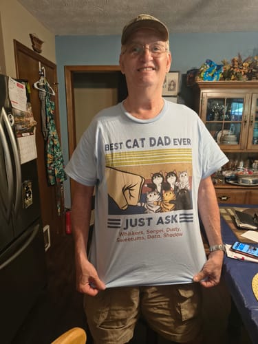 Customer smiling and holding out their light blue personalized "Best Cat Dad Ever" t-shirt with a fist bump graphic and cat names.