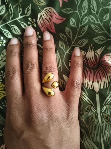 A reviewer's hand wearing the bold, gold Sirena Ring against a green floral background.