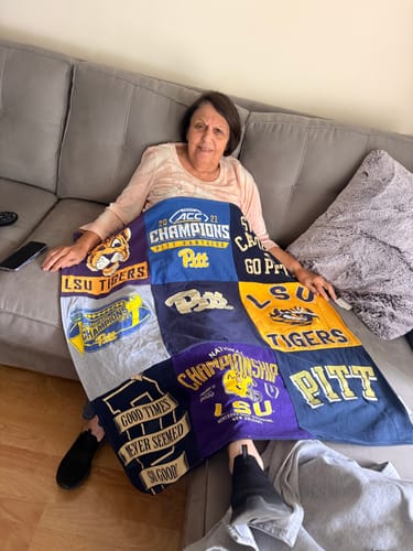 Customer on a couch covered by a colorful Project Repat T-Shirt Quilt with LSU and Pitt logos.