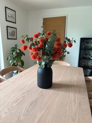 Customer's Umber and Green Faux Arrangement in a black vase, shown on a wooden dining table.