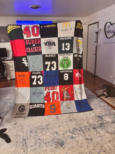 Customer displays a large Project Repat T-Shirt Quilt made of colorful sports and graphic shirts in a living room.