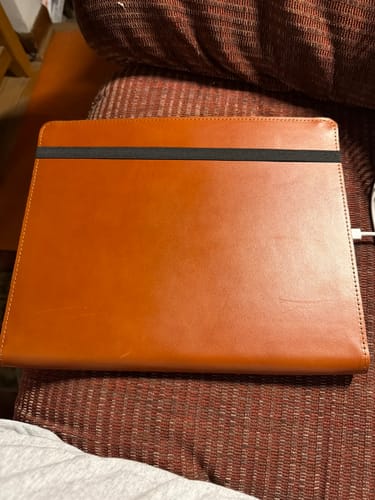 Customer's brown leather English Bridle Magic Keyboard Case, closed with a black strap, resting on a couch.