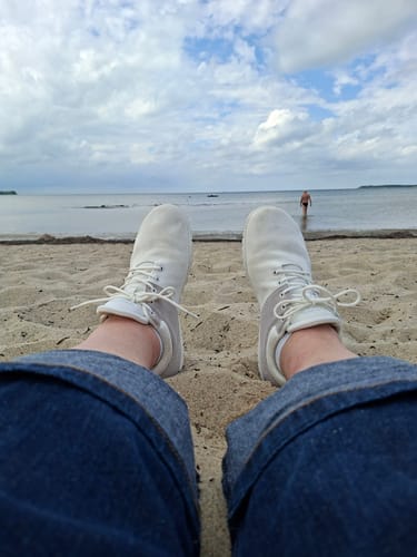 Kunde trägt die weißen Wish I - Barfußschuhe an einem Sandstrand mit Blick auf das Meer.