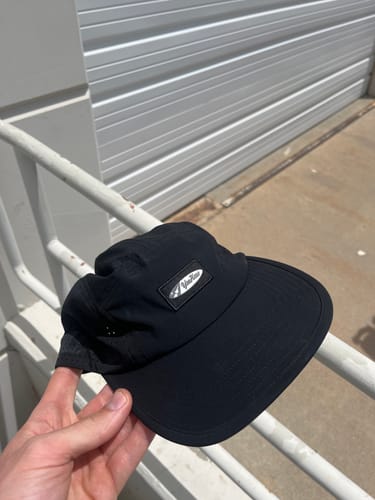 A reviewer's hand holds up the black Big Camp Hat Sport outdoors against a railing.